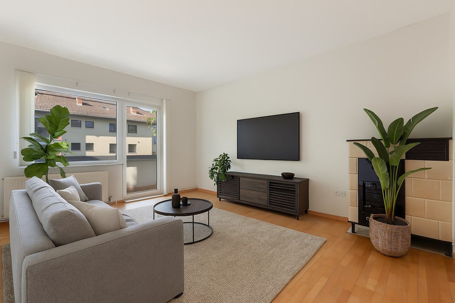Helles Wohnzimmer mit grauem Sofa, rundem Tisch, TV, Pflanzen und großem Fenster.
