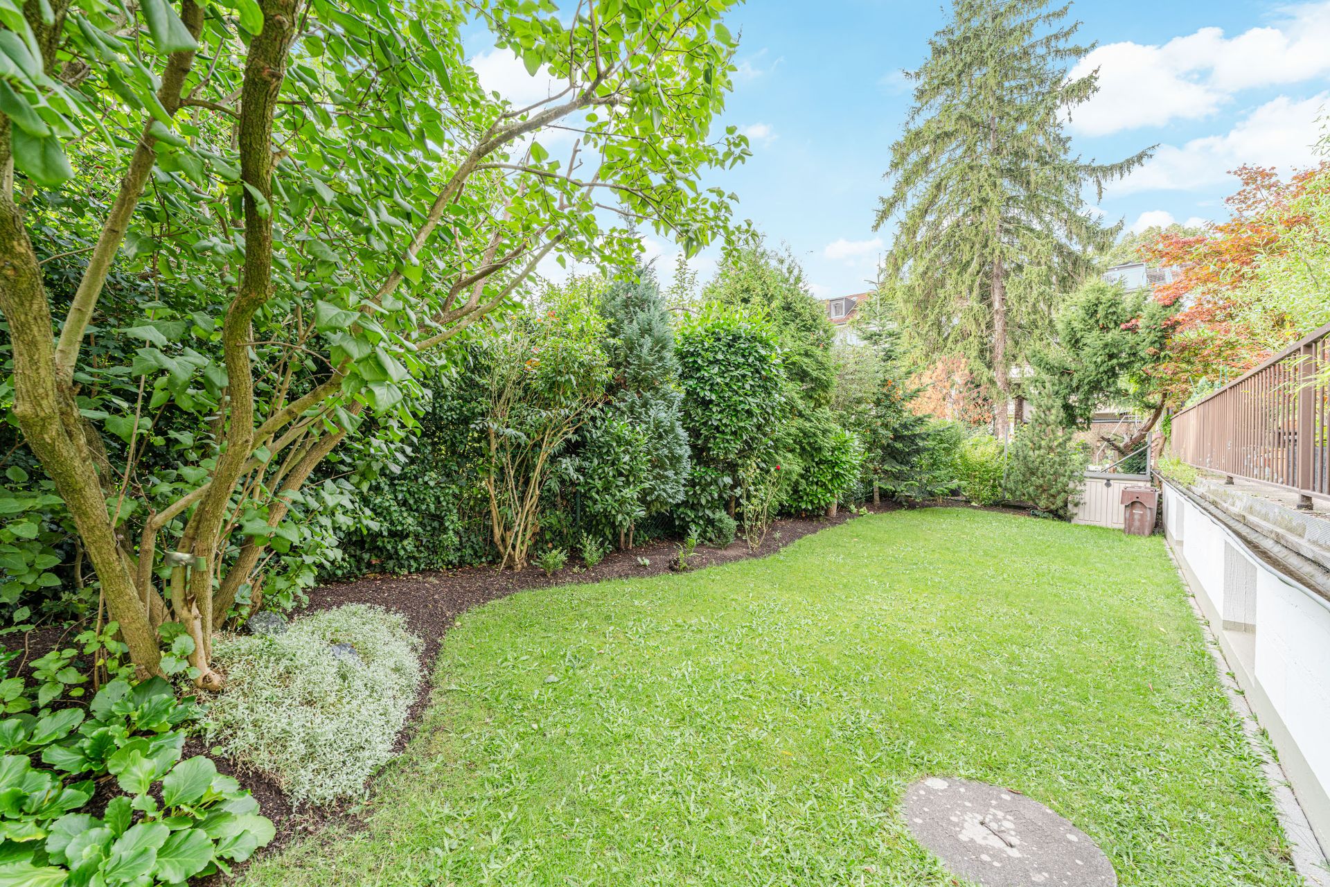 Grüner Garten mit Rasenfläche, Büschen und Bäumen, daneben ein Zaun und einige Gartenhäuser.