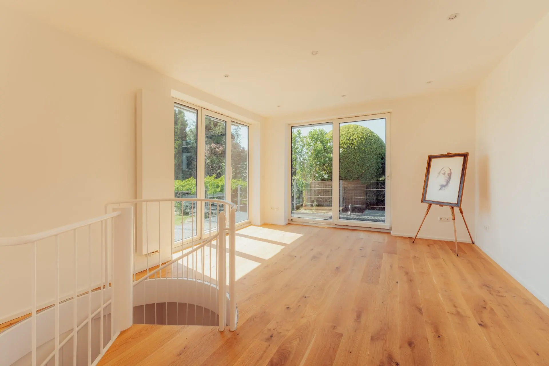 Heller Raum mit Holzboden, großen Fenstern, Zugang zum Balkon und einer runden Treppe.