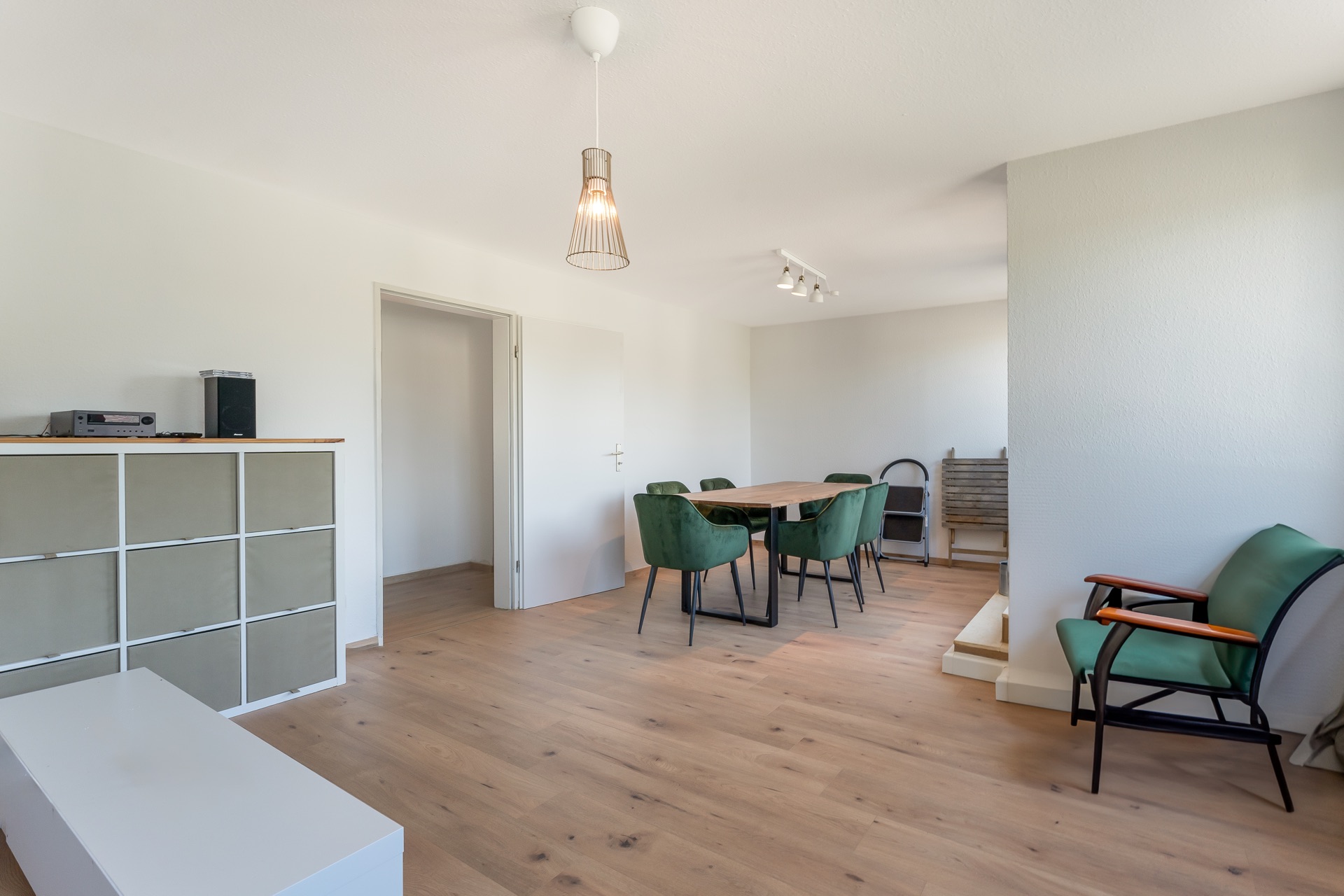Helles Wohnzimmer mit Holzboden, Esstisch, grünen Stühlen und Regal.
