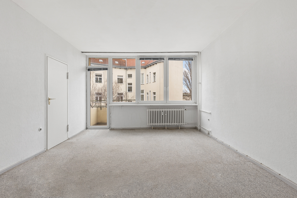 Helles, leeres Zimmer mit Teppichboden, weißen Wänden und großem Fenster mit Tür zum Balkon.