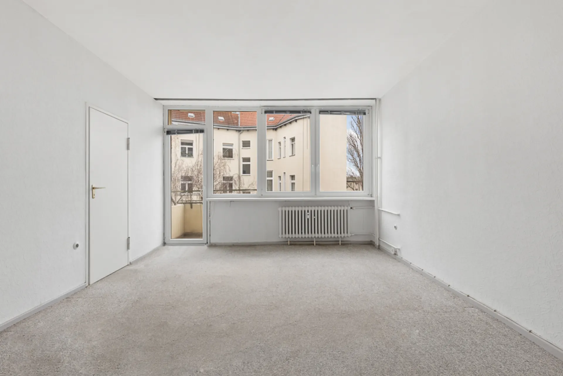 Helles, leeres Zimmer mit Teppichboden, weißen Wänden und großem Fenster mit Tür zum Balkon.