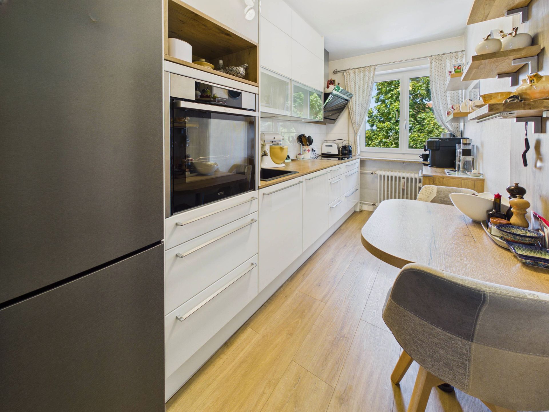 Moderne Küche mit weißen Schränken, Holzboden, Esstisch und großem Fenster.
