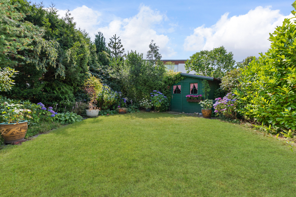 Grüner Garten mit Rasenfläche, Blumenbeeten und einem grünen Gartenschuppen im Hintergrund.