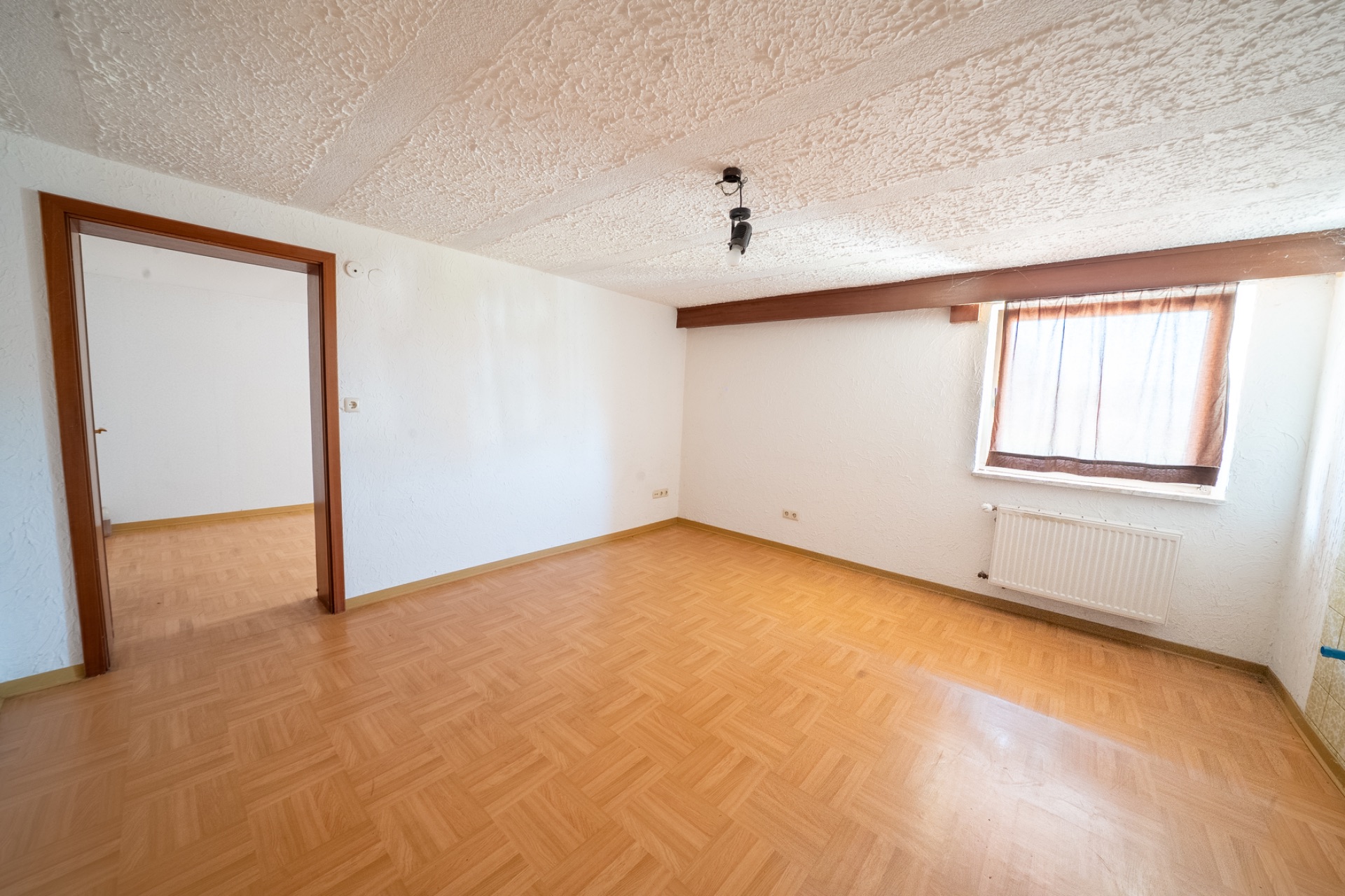Leeres Zimmer mit Parkettboden, Fenster mit Vorhang und Tür zu einem weiteren Raum.