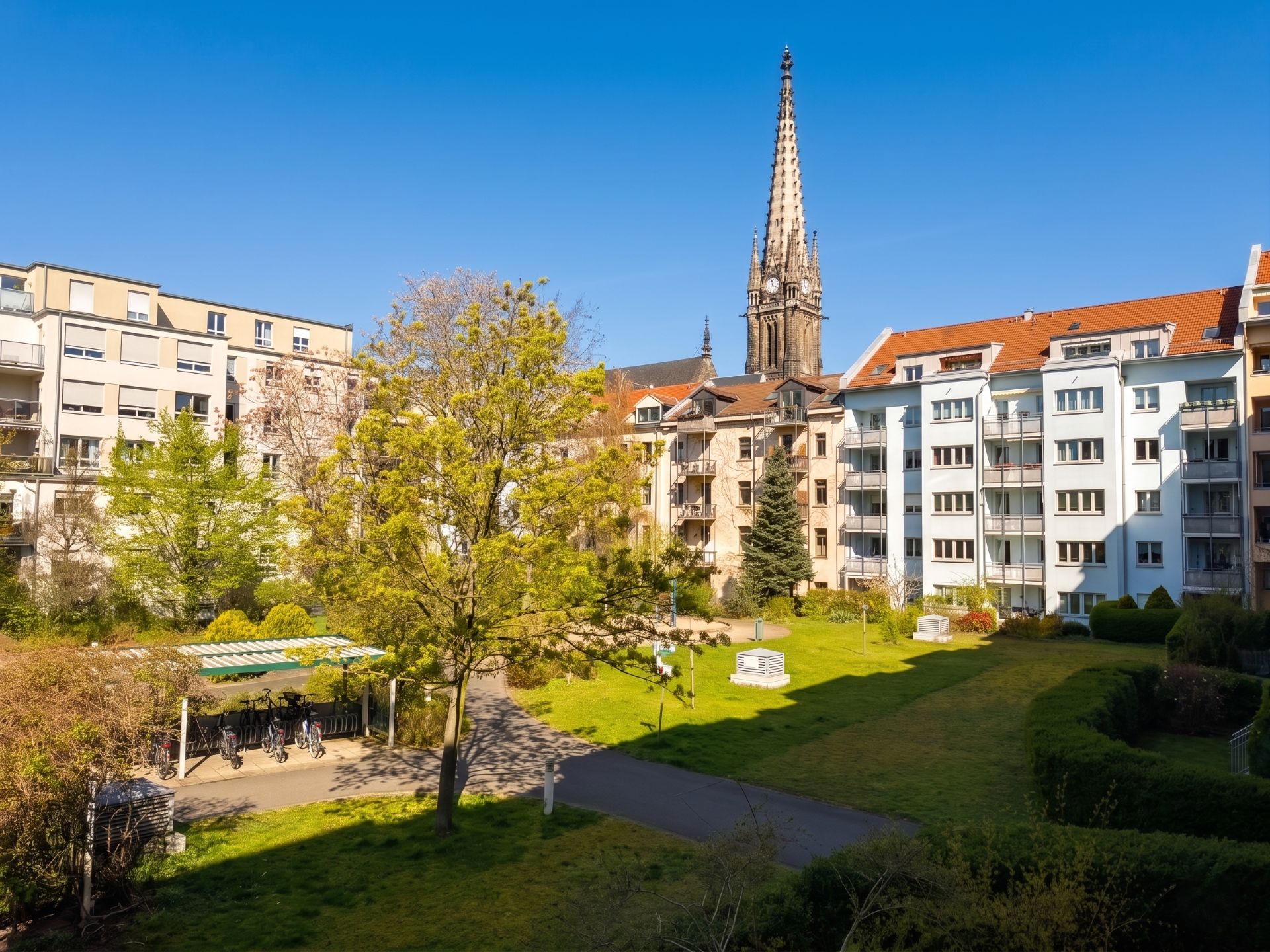Grüne Wohnanlage mit mehrstöckigen Gebäuden und Kirche im Hintergrund bei blauem Himmel.