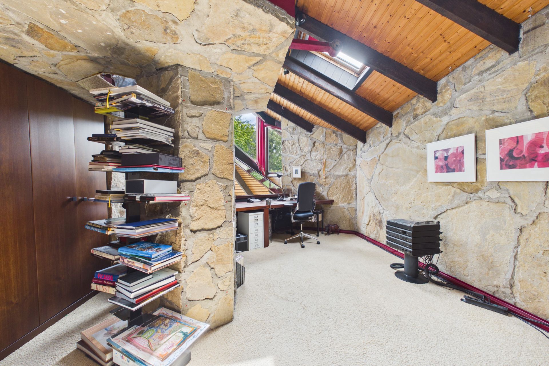 Arbeitszimmer mit Steintapete, Schreibtisch, Bücherregal und schrägem Holzplafond.