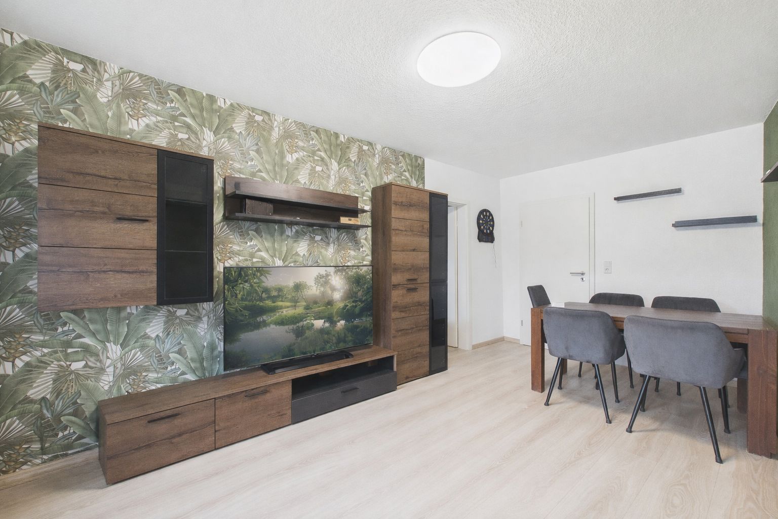 Wohnzimmer mit Holzmöbeln, TV, Esstisch und vier grauen Stühlen vor gemusterter Tapete.