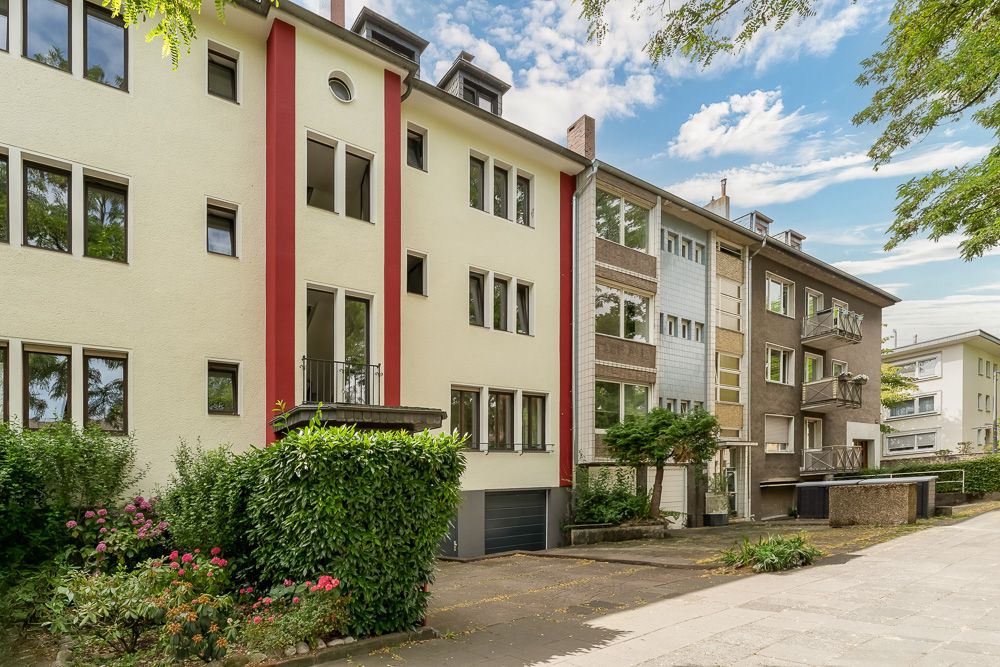 Mehrfamilienhaus mit mehreren Fenstern, Balkonen und grüner Bepflanzung vor dem Gebäude.