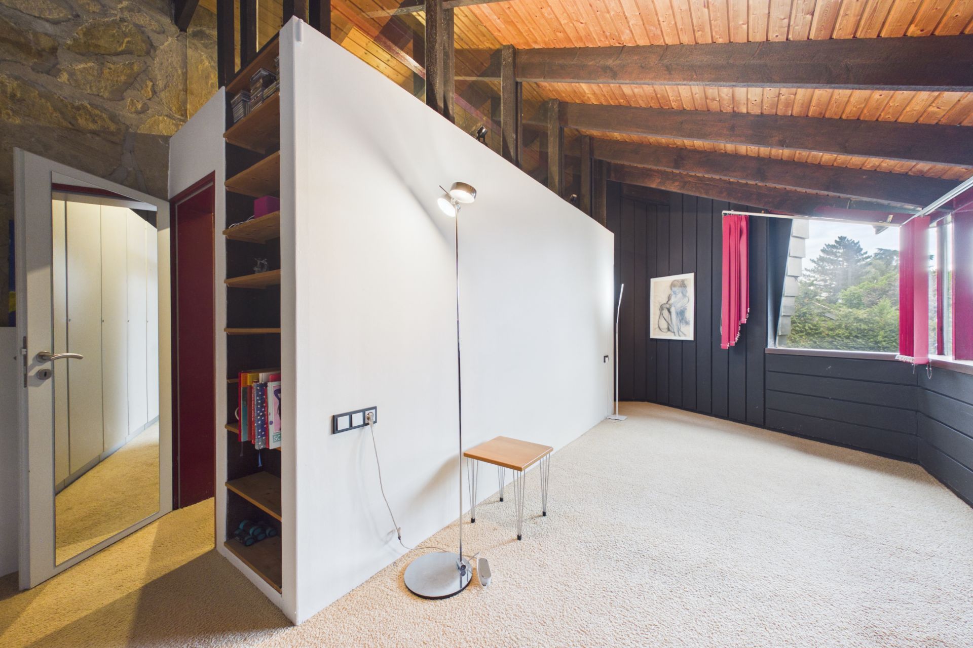 Helles Zimmer mit Holzbalkendecke, Teppichboden, Wandregal, Stehlampe, Hocker und großen Fenstern mit roten Vorhängen.