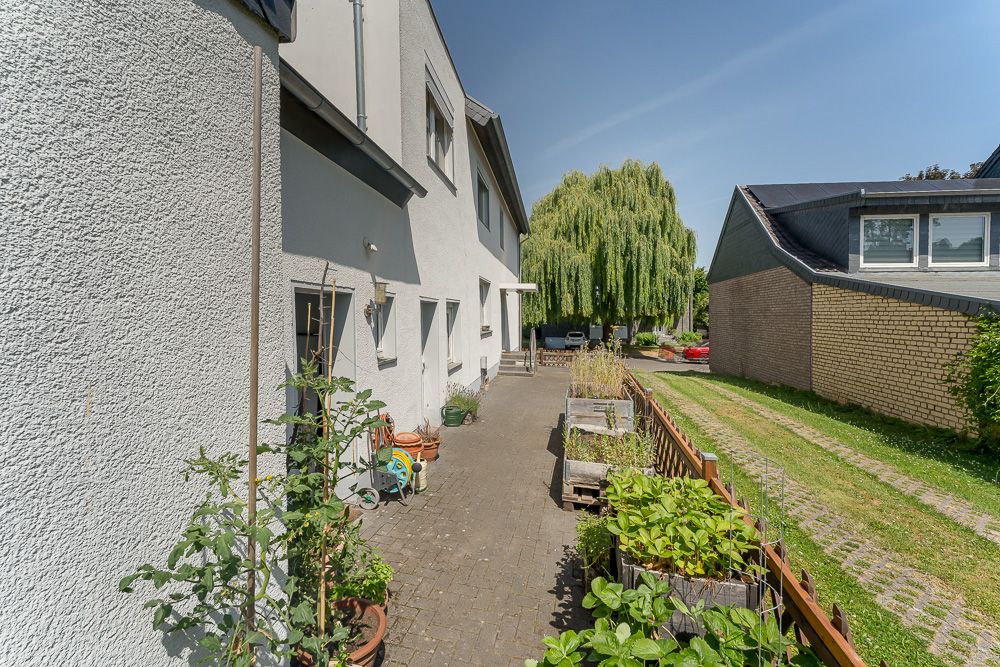 Gepflasterter Weg neben einem Wohnhaus mit Pflanzen und Beeten, daneben eine Wiese.