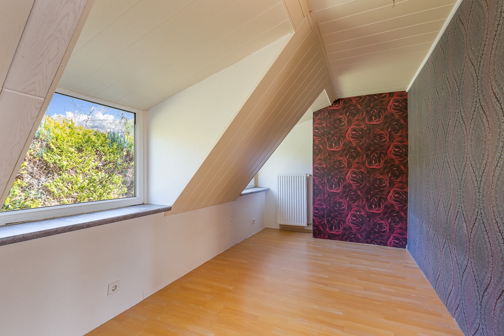 Heller Raum mit schräger Decke, großem Fenster, Holzfußboden und gemusterter Tapete.