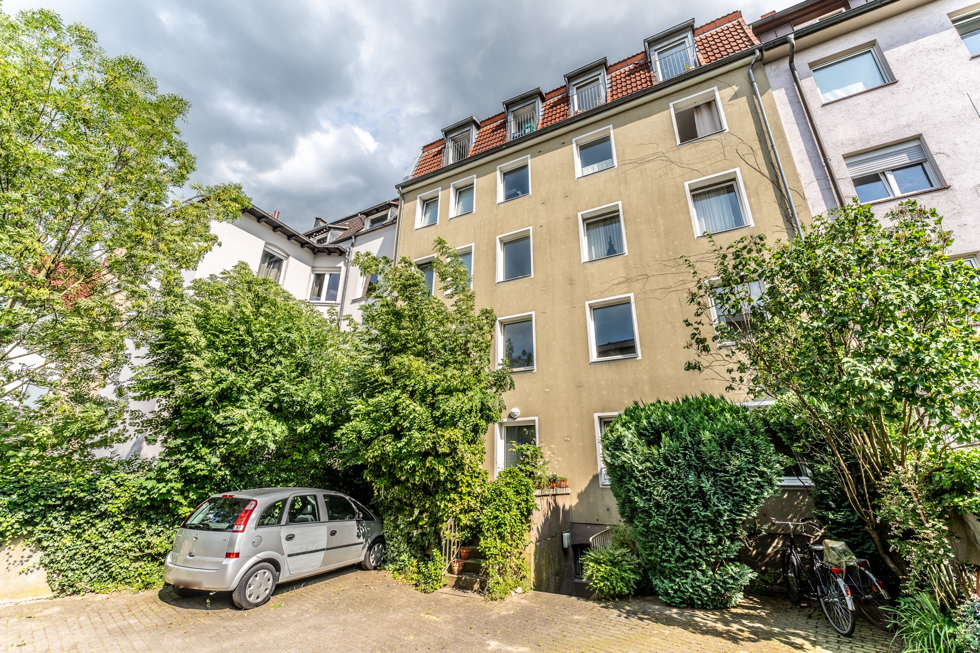 Mehrstöckiges Wohnhaus mit vielen Fenstern, umgeben von Bäumen, mit geparktem Auto und Fahrrädern vorne.