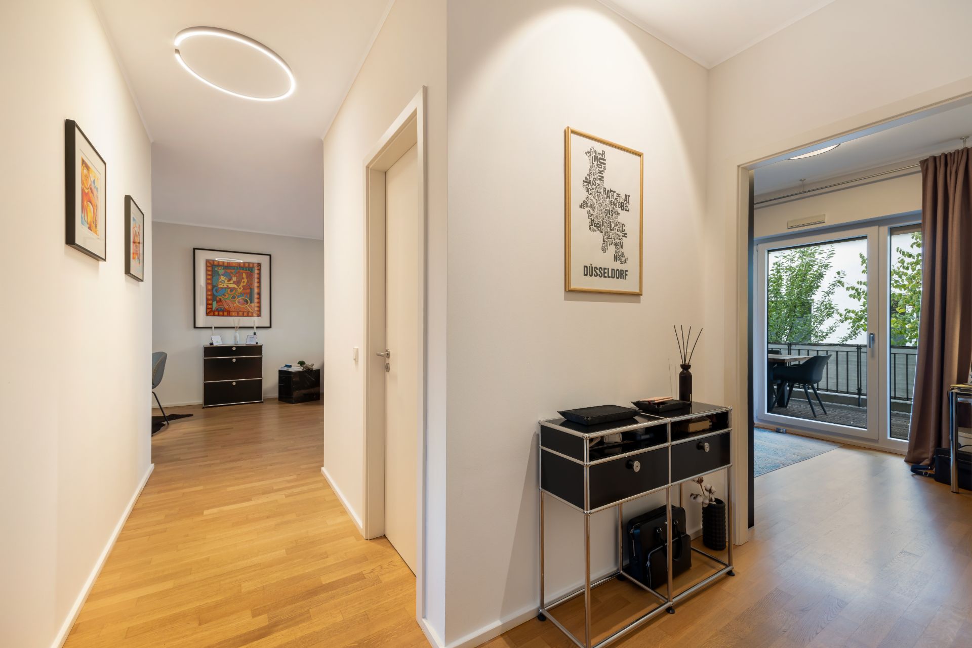 Heller Flur mit Holzboden, Wandbildern, Kommode und Blick in ein Zimmer mit Balkon.