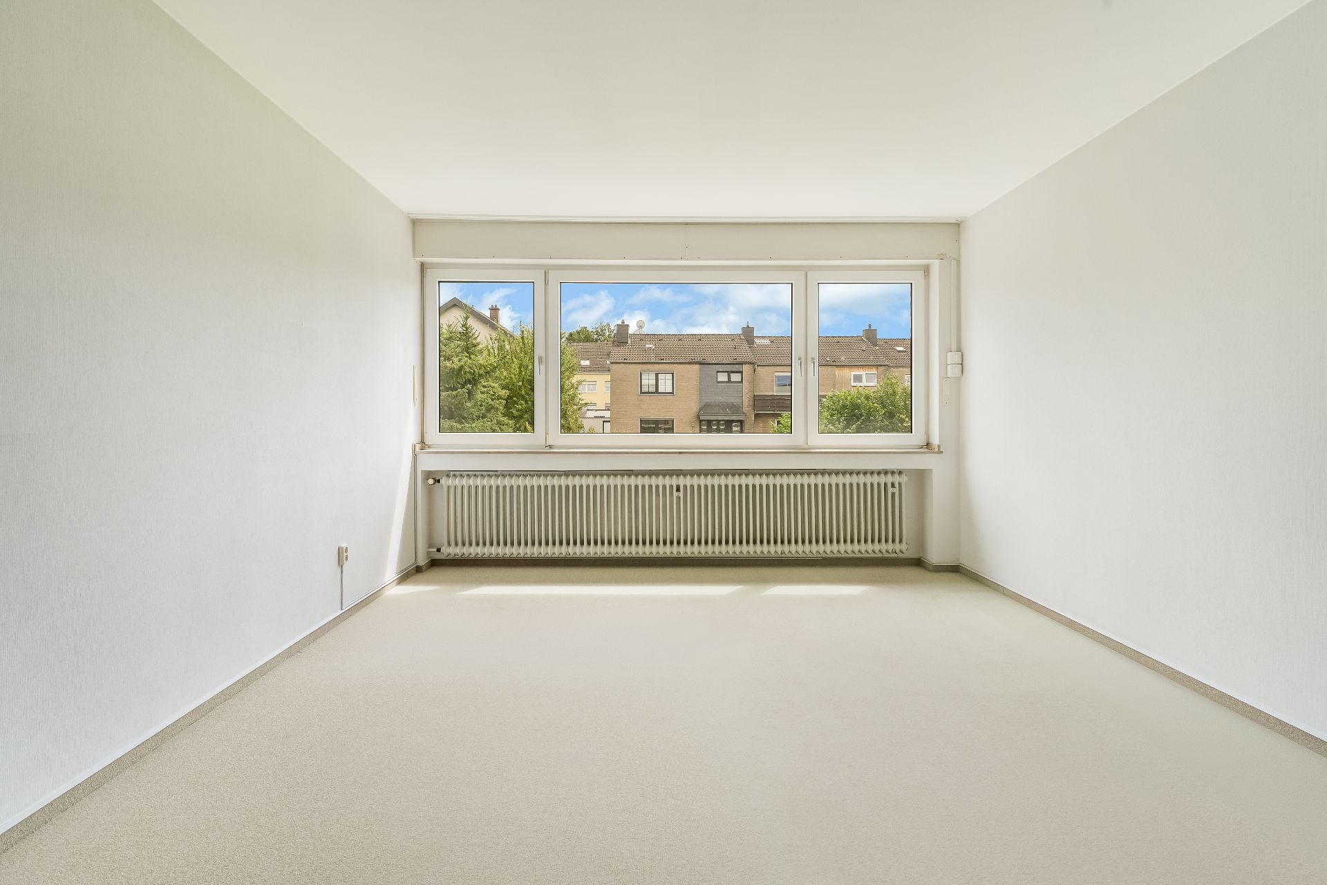 Leerer, heller Raum mit großem Fenster, Heizung darunter und Blick auf Häuser gegenüber.