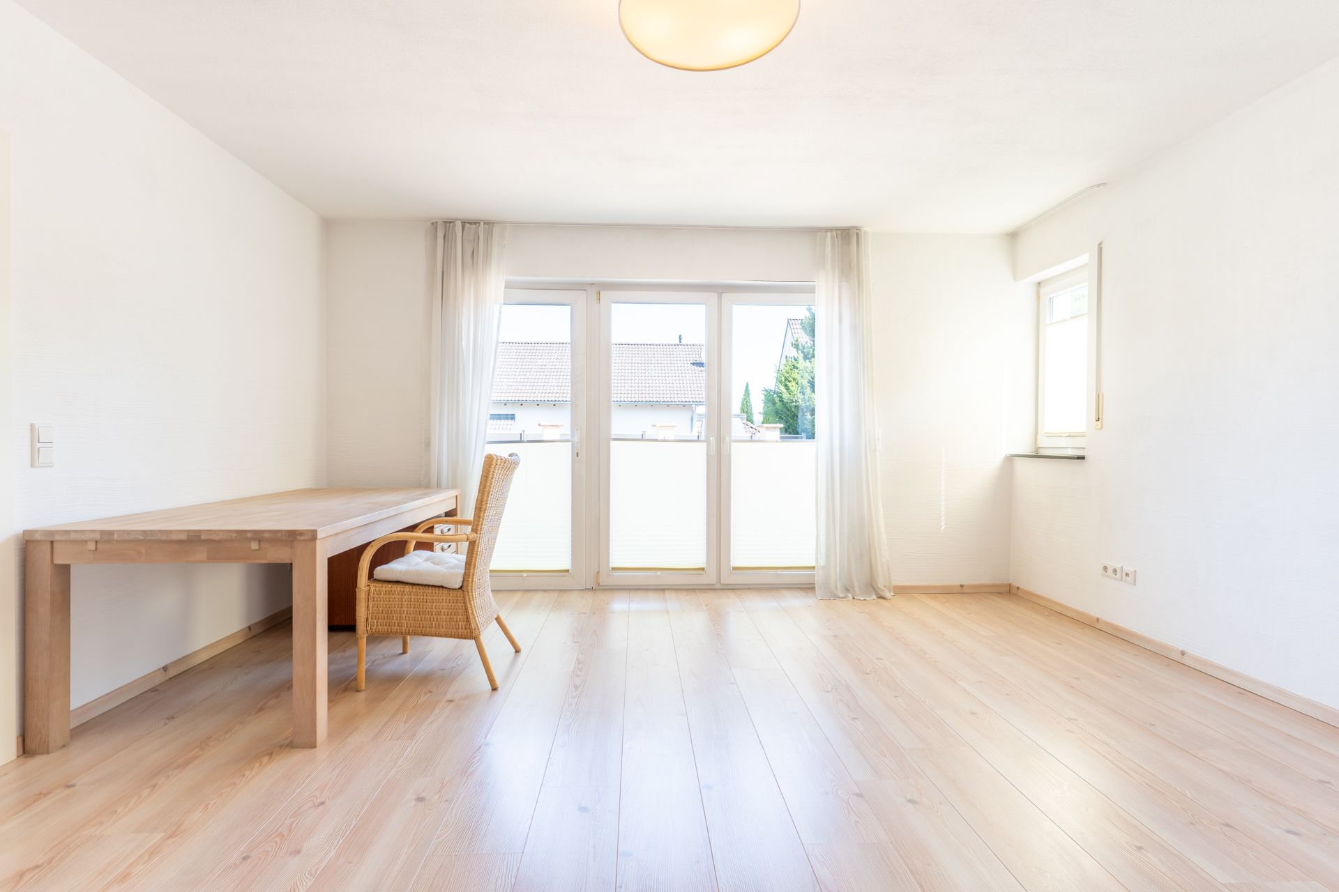 Helles Zimmer mit großem Fenster, Holztisch und Stuhl auf hellem Holzfußboden.