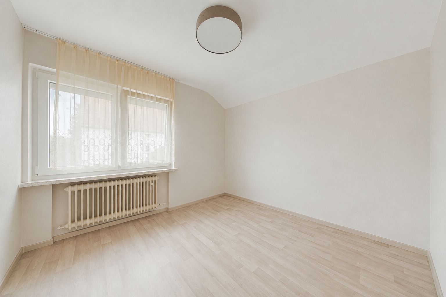 Leeres Zimmer mit hellen Wänden, großem Fenster, Gardine und Holzfußboden.