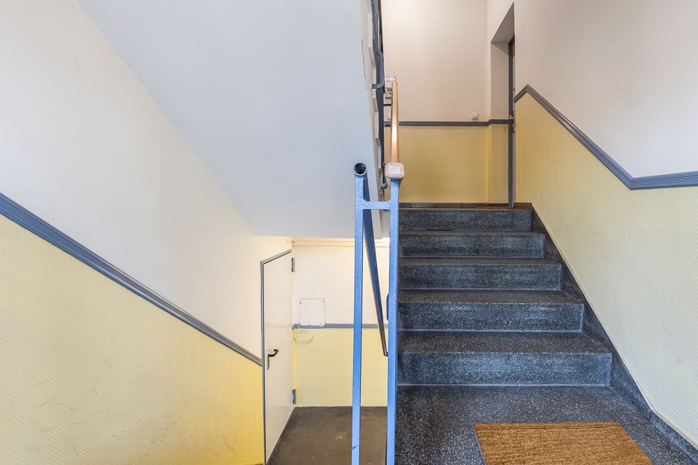 Treppenhaus mit grauen Stufen, gelben Wänden und einer Eingangstür unten.