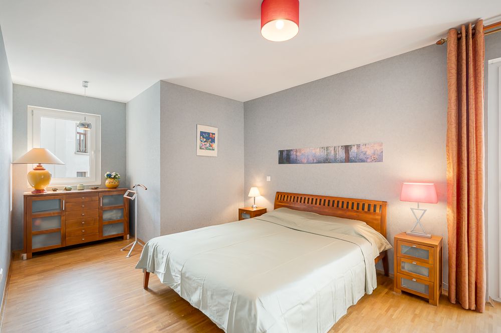Schlafzimmer mit Doppelbett, Nachttischen, Kommode und großem Fenster mit Vorhang.