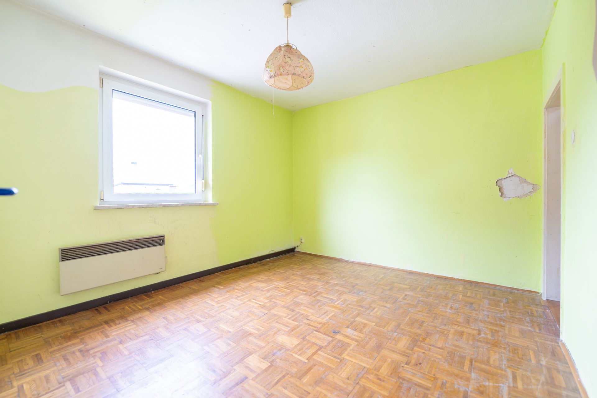 Leerer Raum mit Parkettboden, grünen Wänden, Fenster und Heizkörper, beschädigte Stelle an der Wand.