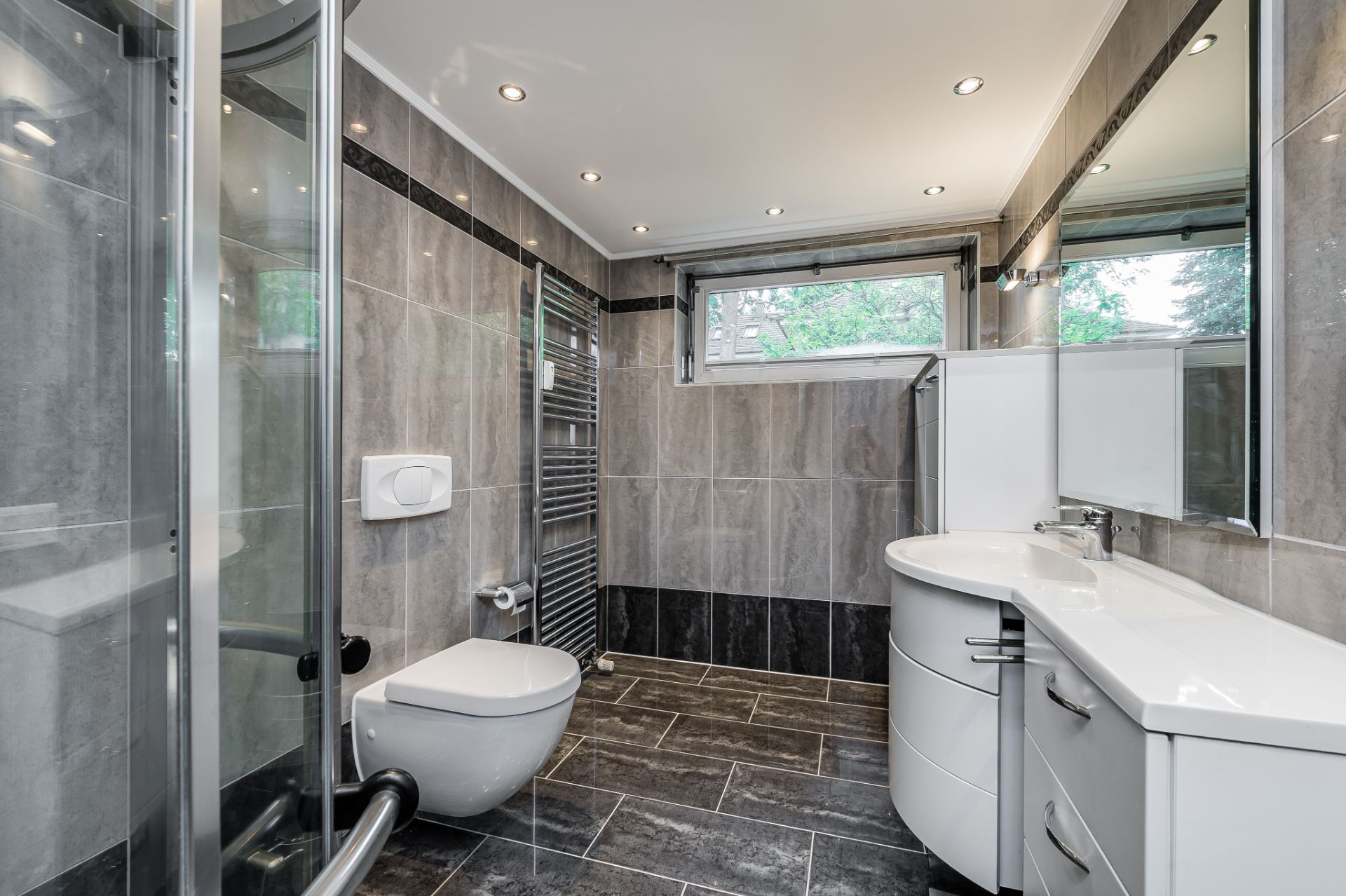 Modernes Badezimmer mit Dusche, WC, Waschtisch und großem Spiegel, graue Fliesen an Wänden und Boden.