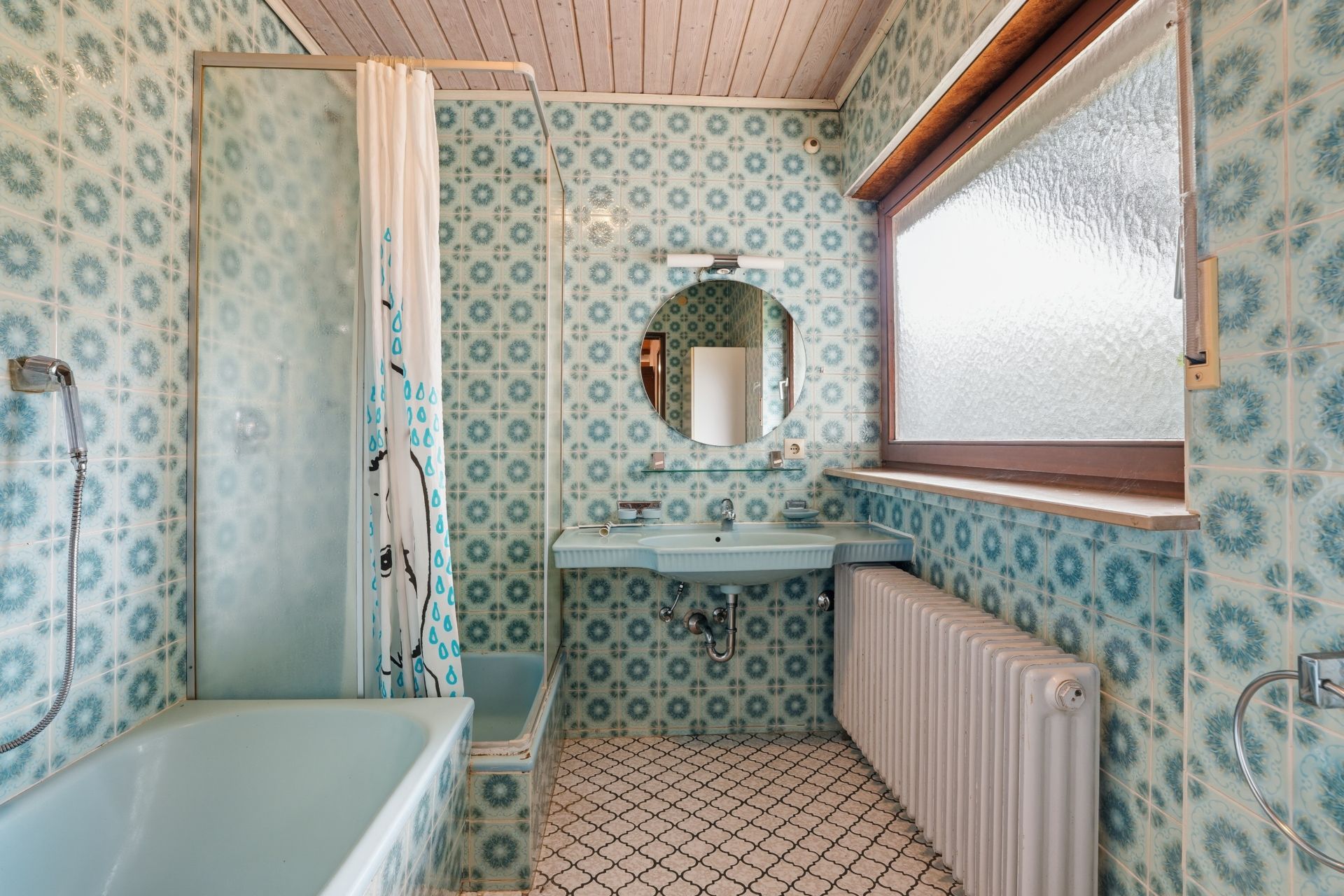 Badezimmer mit gemusterten blauen Fliesen, Badewanne, Waschbecken, Rundspiegel und Fenster.