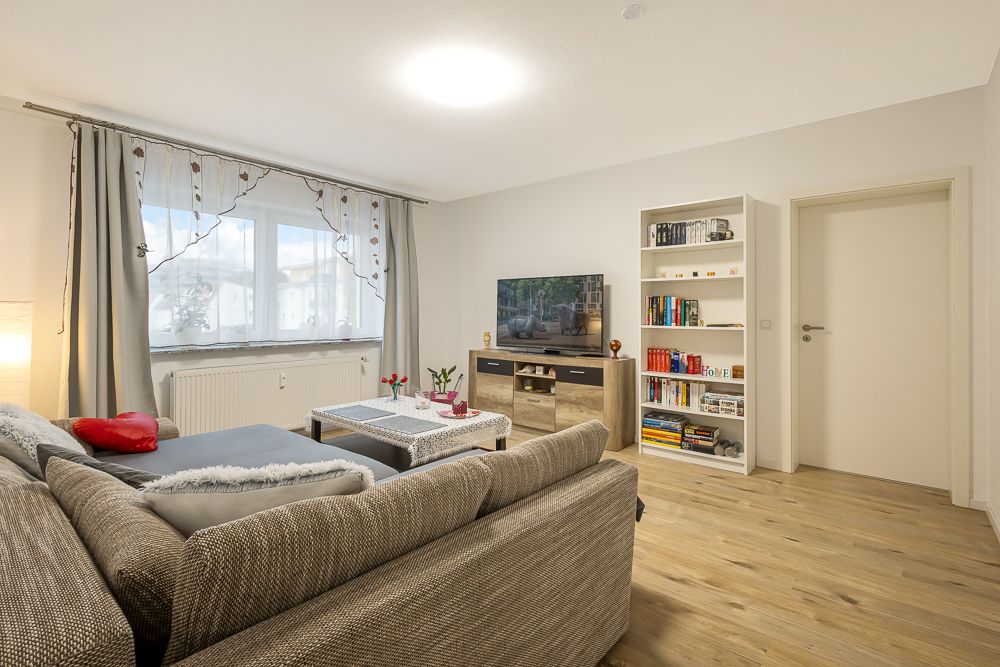 Wohnzimmer mit grauem Sofa, Couchtisch, Fernseher, Bücherregal und großem Fenster mit Gardinen.