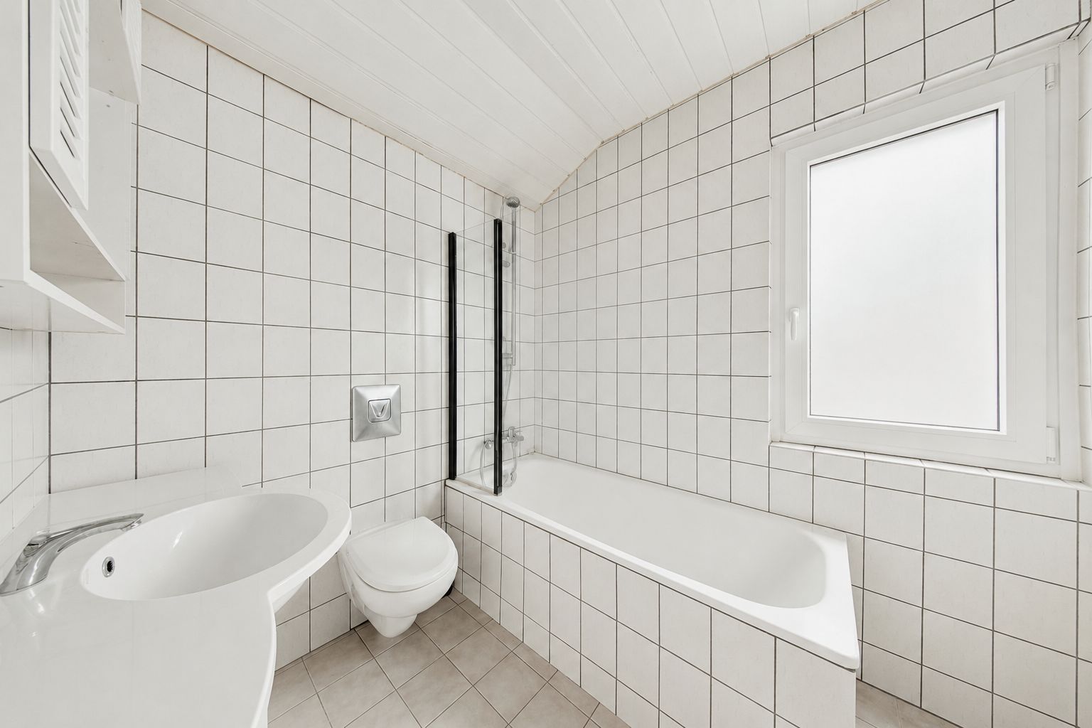 Helles Badezimmer mit Badewanne, Waschbecken, WC und Fenster, alle Wände sind weiß gefliest.