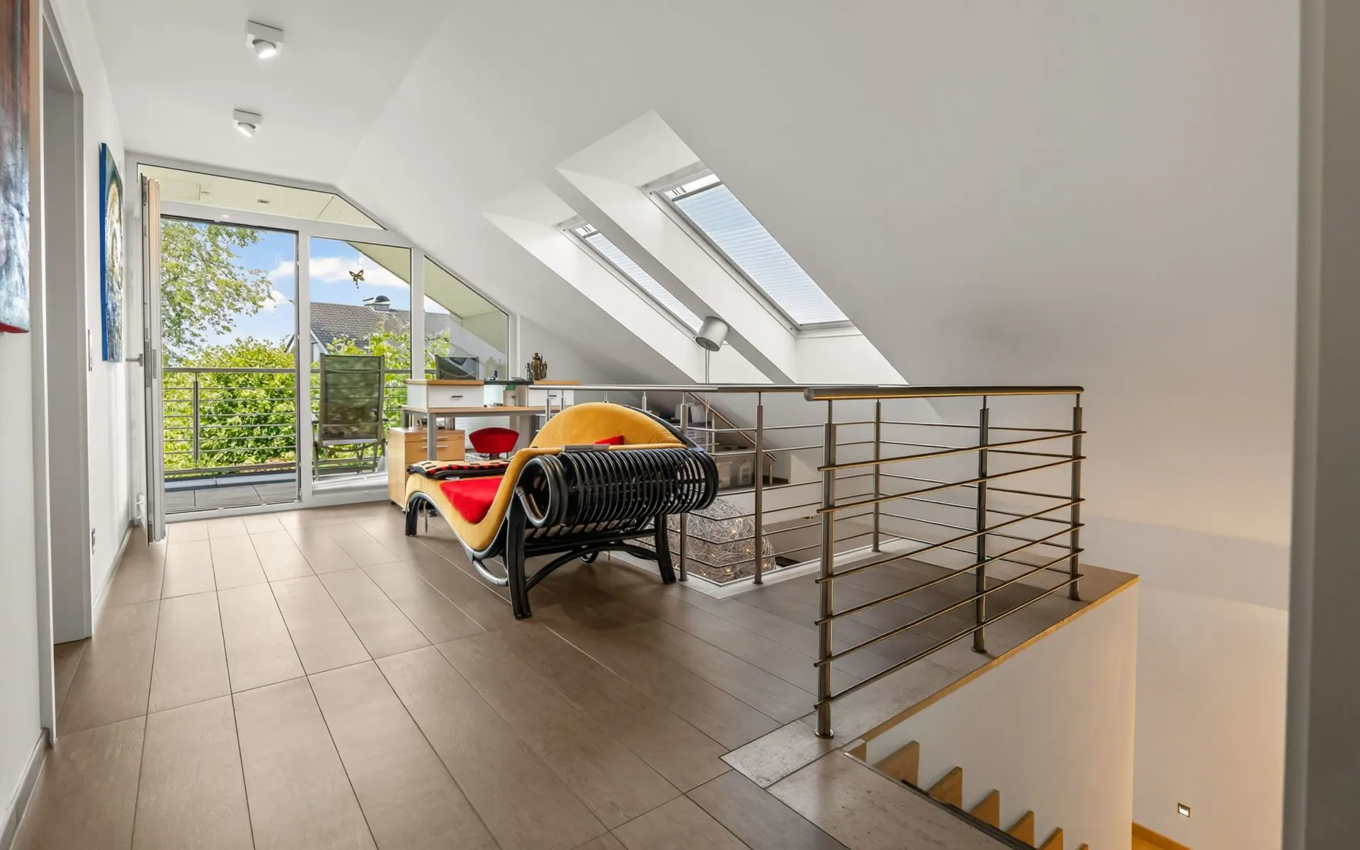 Heller Dachbodenbereich mit Sofa, Schreibtisch, Fenstern, Zugang zu Balkon und Treppenhaus.
