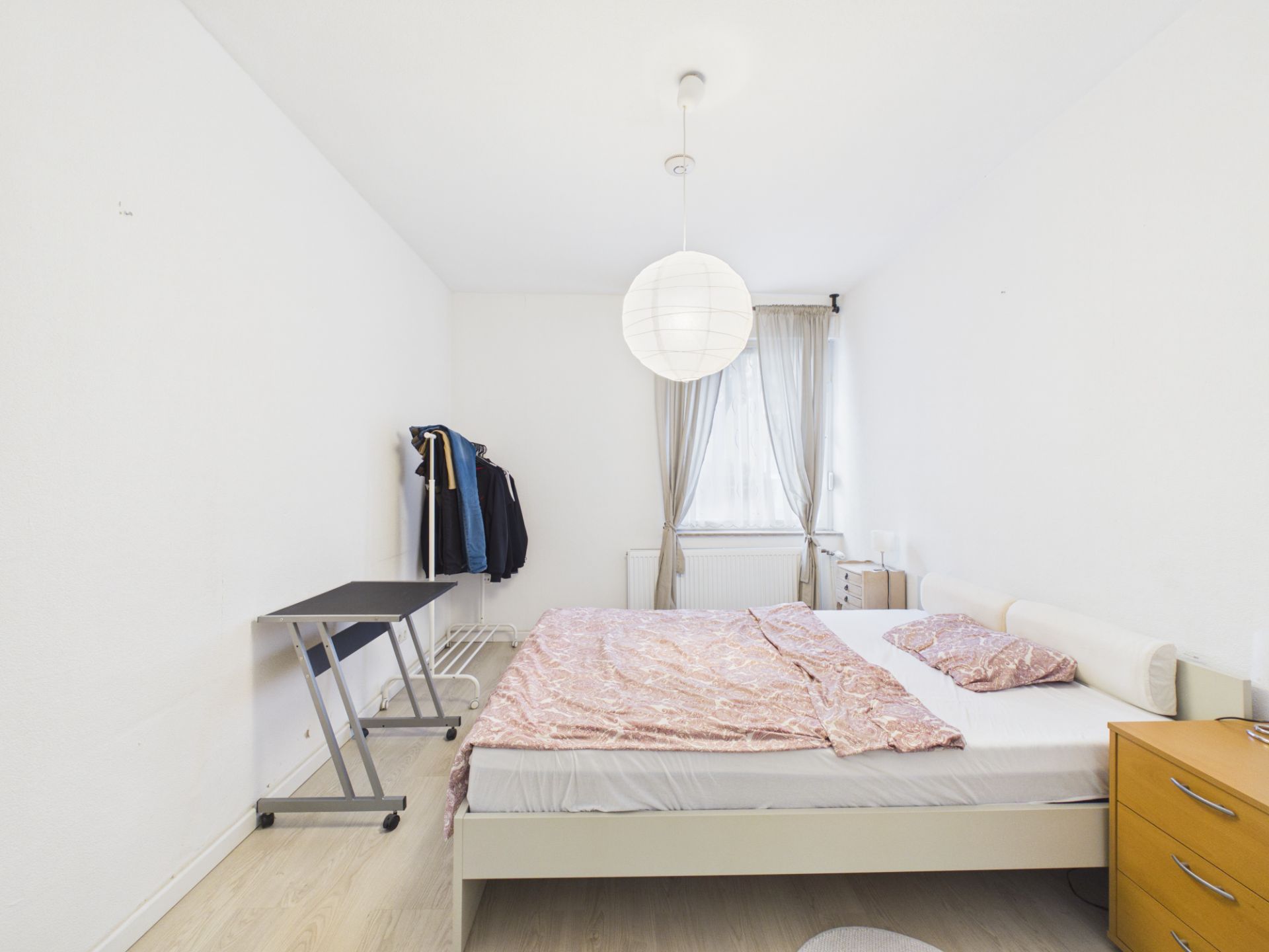 Helles Schlafzimmer mit Doppelbett, Schreibtisch, Kommode und Fenster mit Vorhängen.
