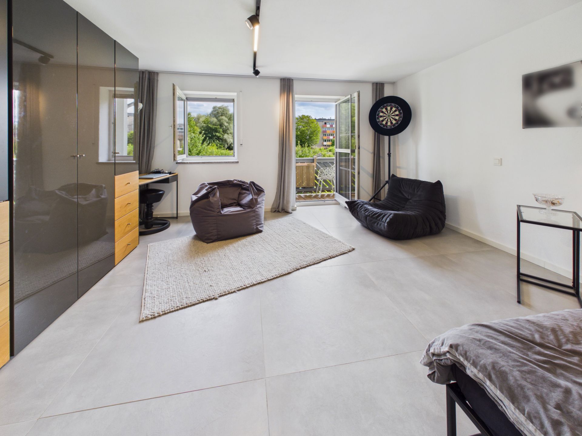 Modernes Zimmer mit Sitzsäcken, Schrank, Schreibtisch, Teppich und offenem Fenster zum Balkon.