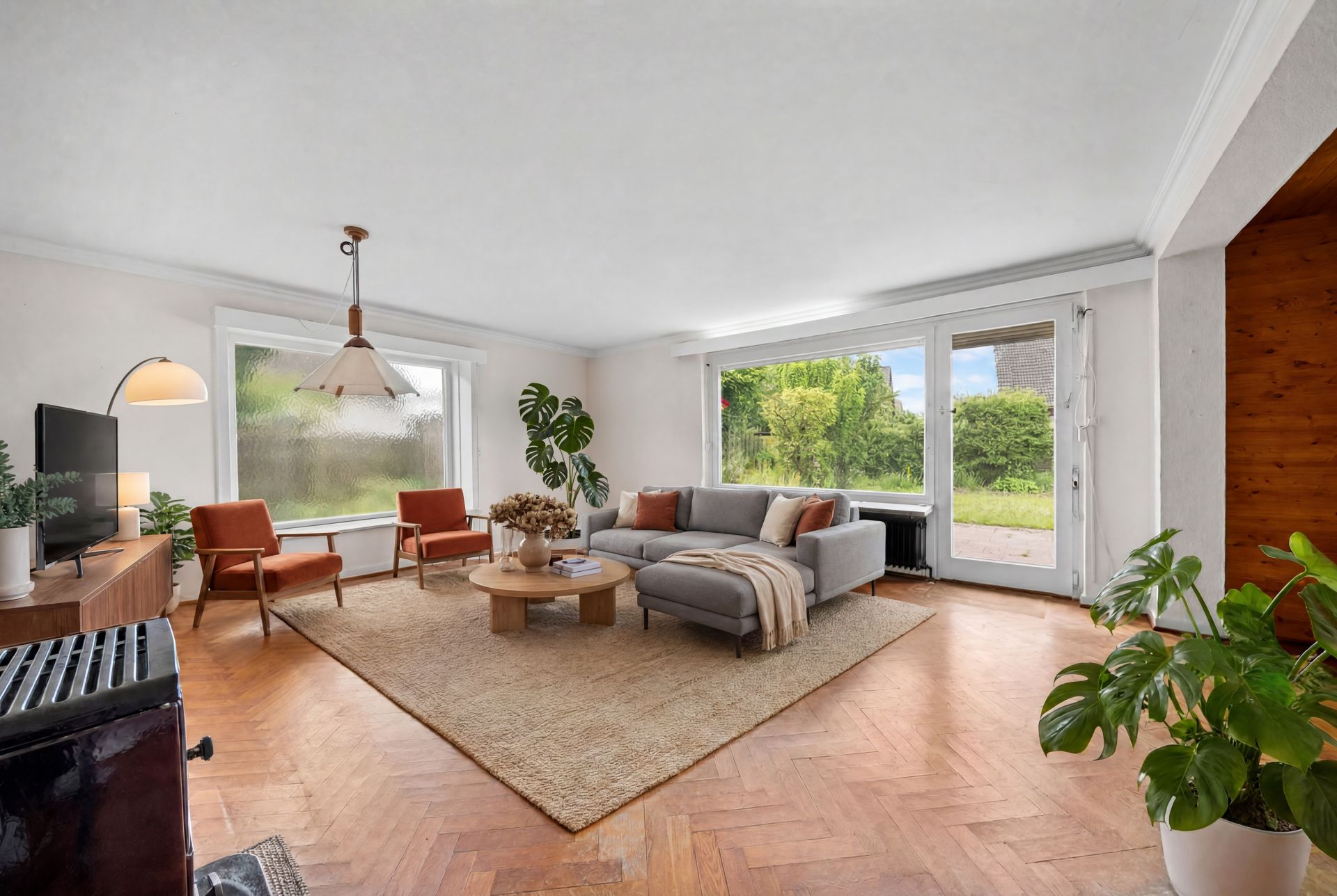 Helles Wohnzimmer mit Sofa, zwei Sesseln, Couchtisch, großem Fenster und Zimmerpflanzen.