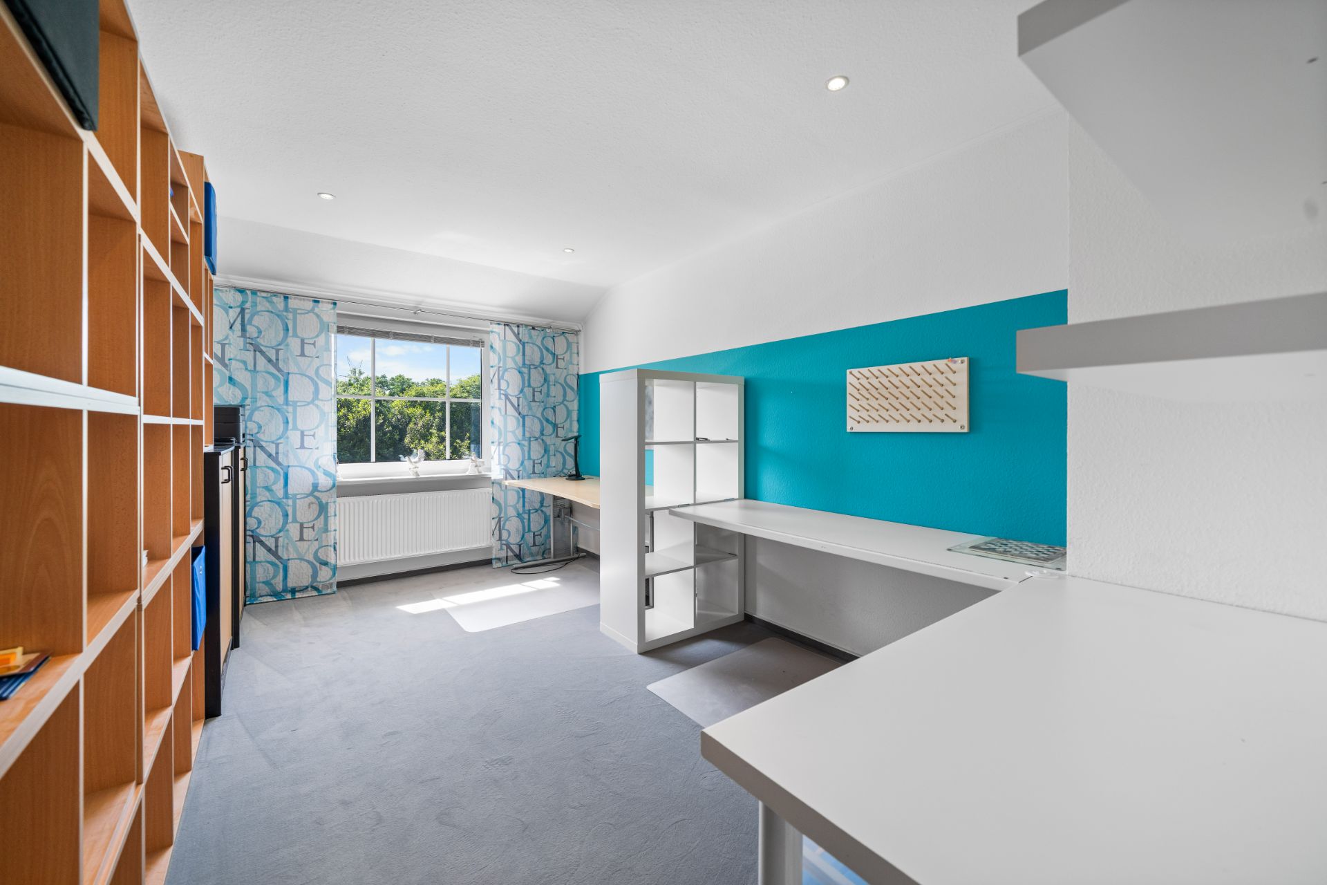 Helles Arbeitszimmer mit großem Fenster, Regalen und blau gestrichener Wand.