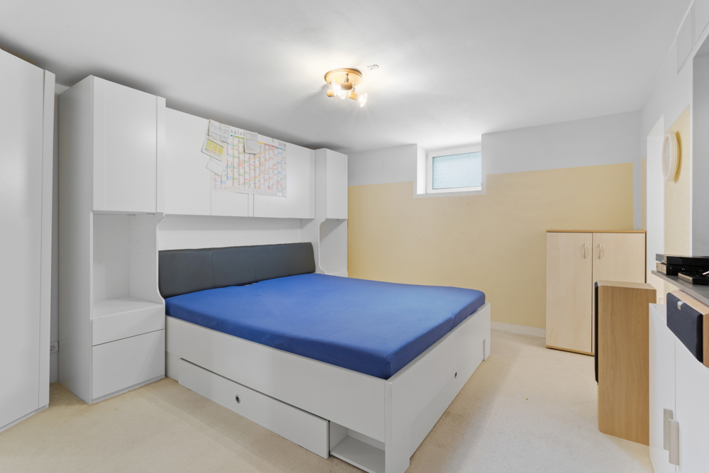 Helles Schlafzimmer mit blauem Bett, weißen Schränken und kleinem Fenster an der Wand.