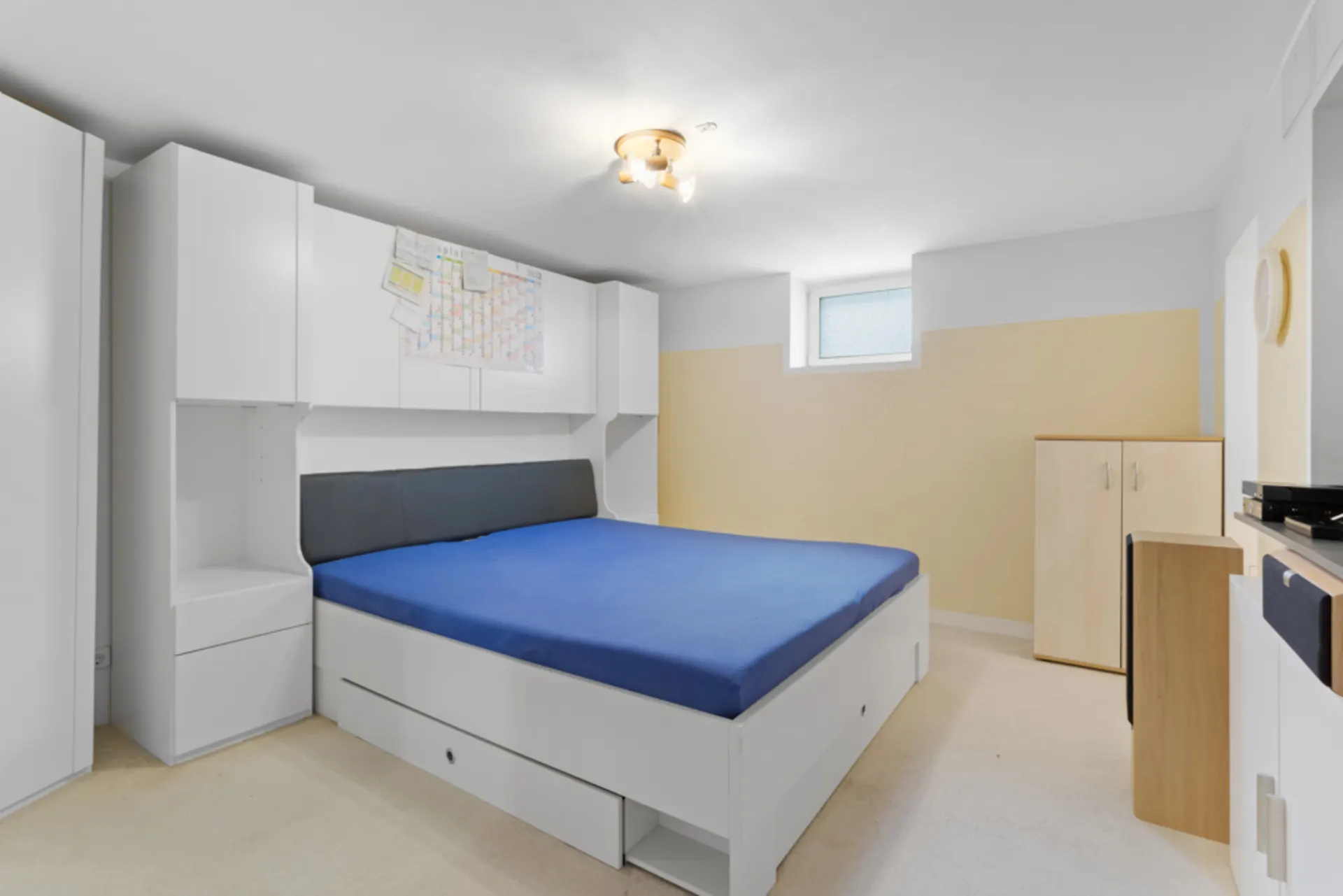 Helles Schlafzimmer mit blauem Bett, weißen Schränken und kleinem Fenster an der Wand.