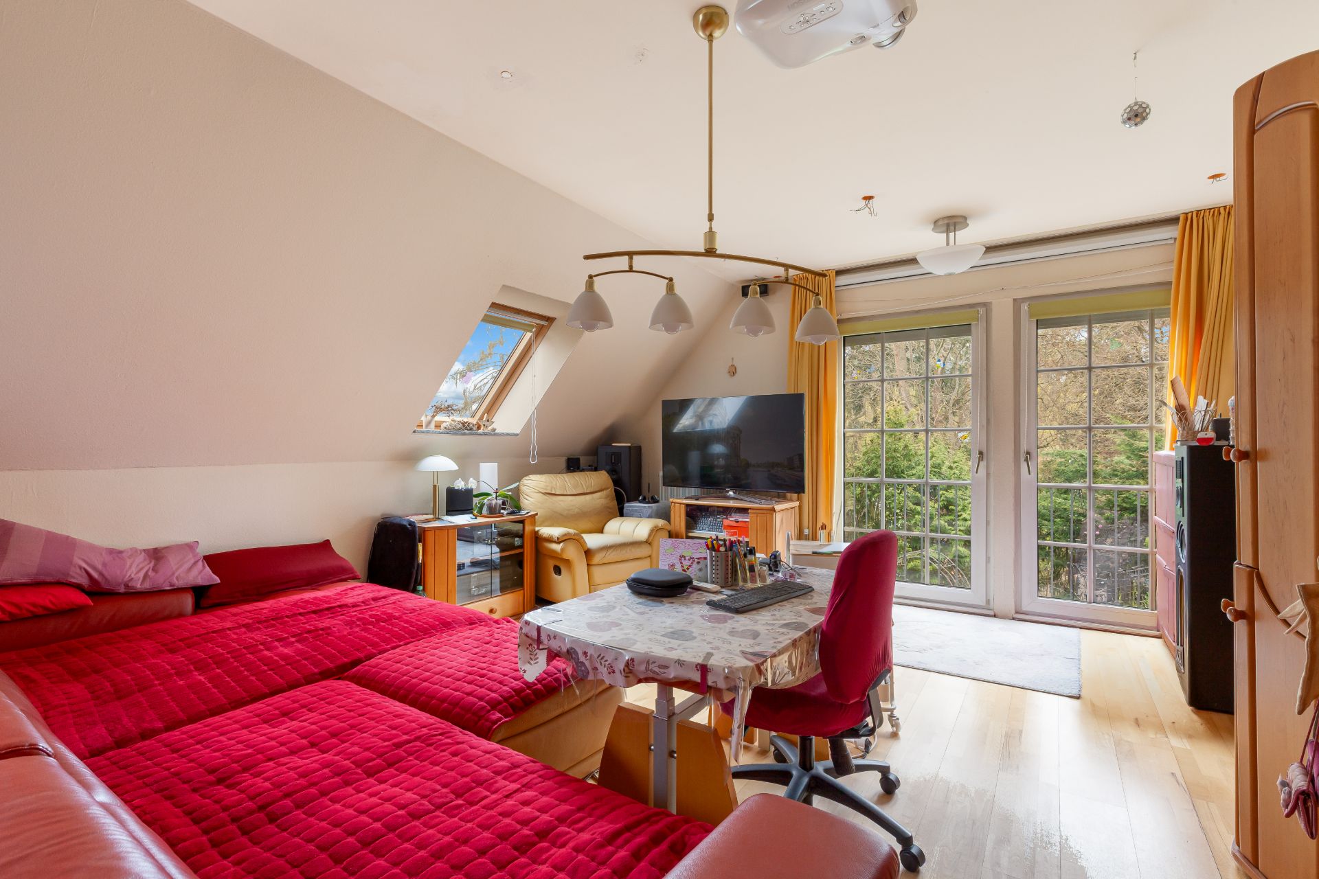 Helles Zimmer mit rotem Bett, Tisch, Sessel, Fernseher und Fenstertüren zum Garten.