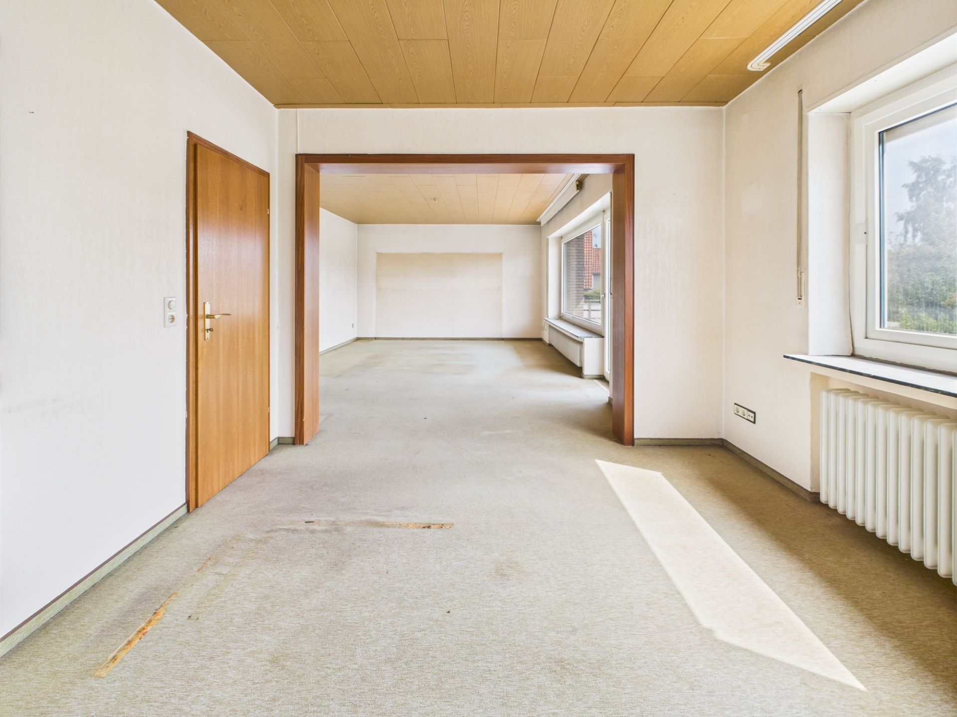 Leerer Raum mit Teppichboden, Holztür, großen Fenstern und Heizkörper unter dem Fenster.