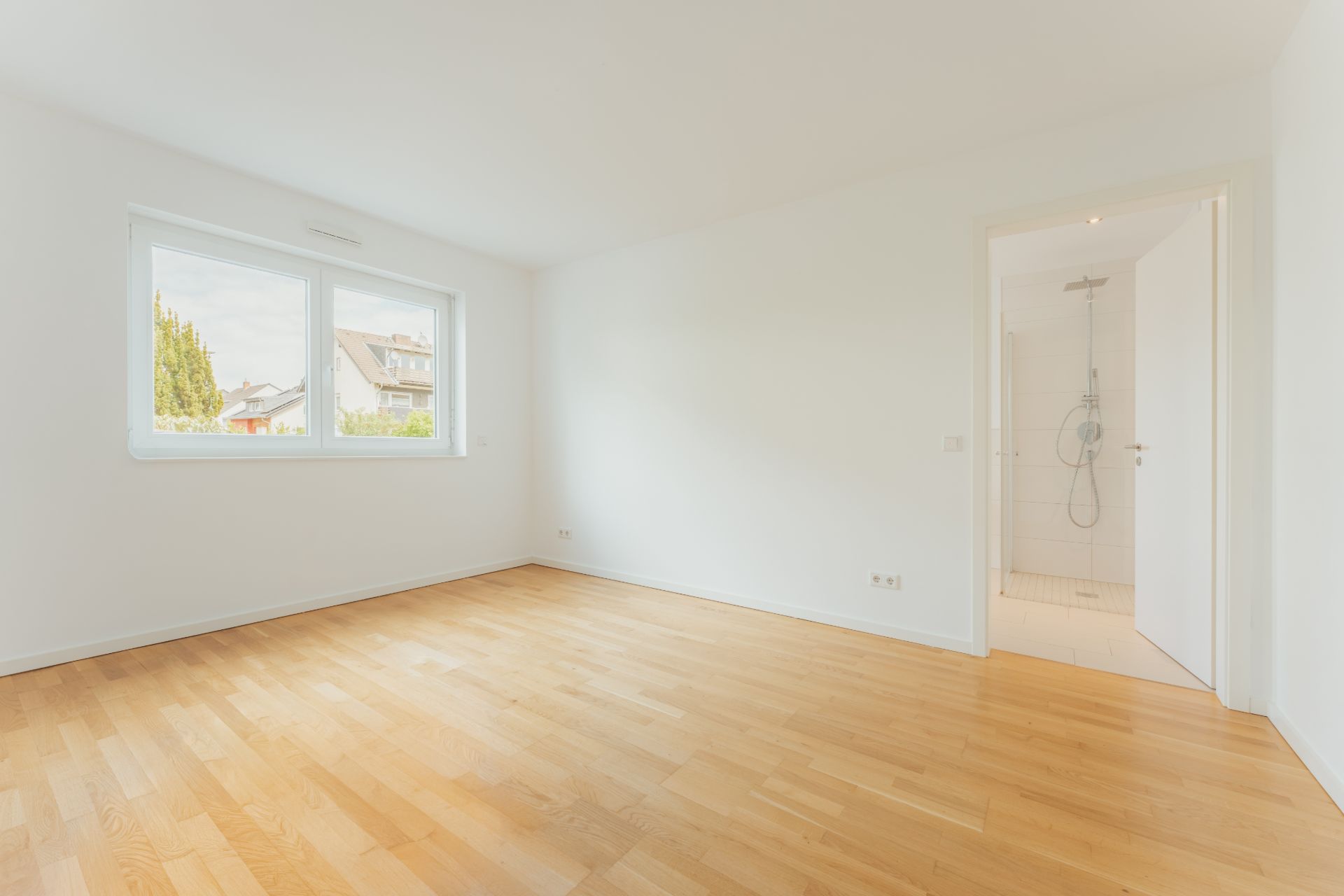 Helles, leeres Zimmer mit Holzfußboden, Fenster und Zugang zu einem Badezimmer mit Dusche.