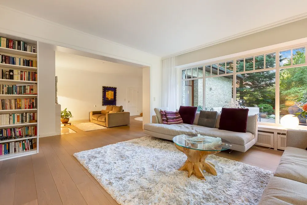 Helles Wohnzimmer mit großem Fenster, Sofa, flauschigem Teppich, Glastisch und Bücherregal.