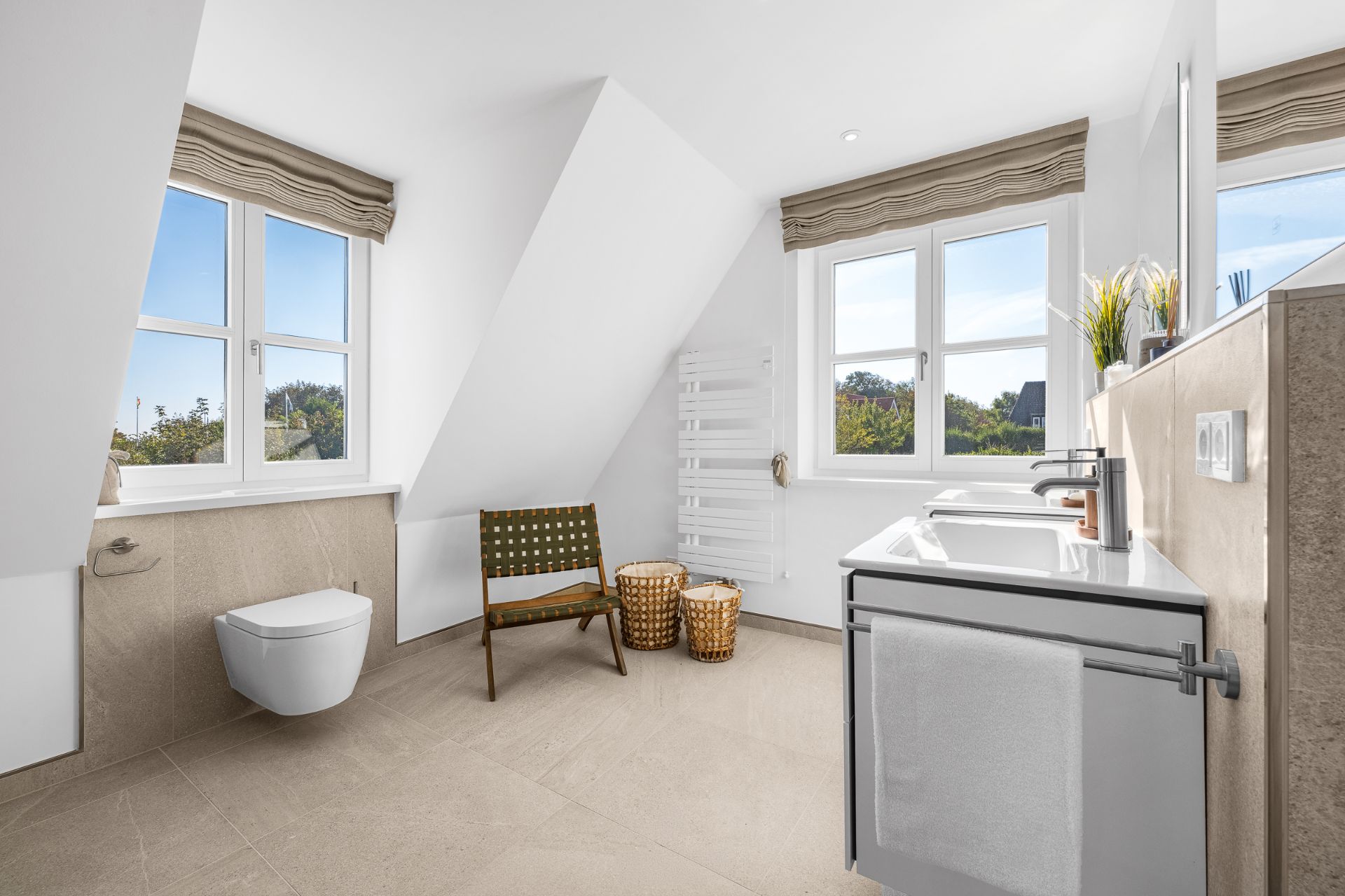 Helles Badezimmer mit WC, Waschbecken, Stuhl, zwei Körben und Fenstern.
