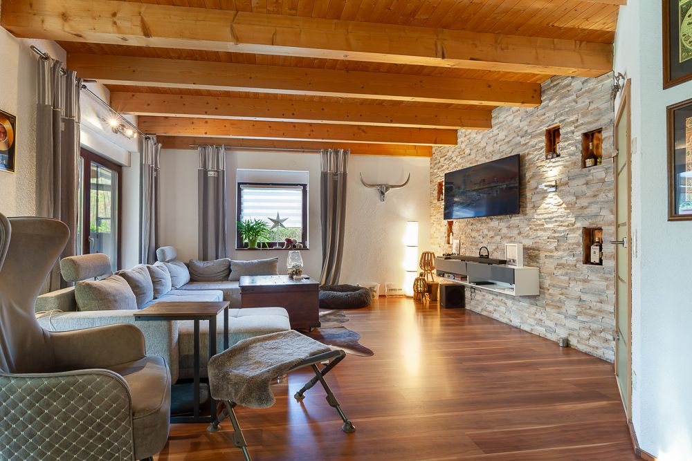 Modernes Wohnzimmer mit Holzbalkendecke, Sofa, Sessel, Fernseher an Steinwand und Holzfußboden.