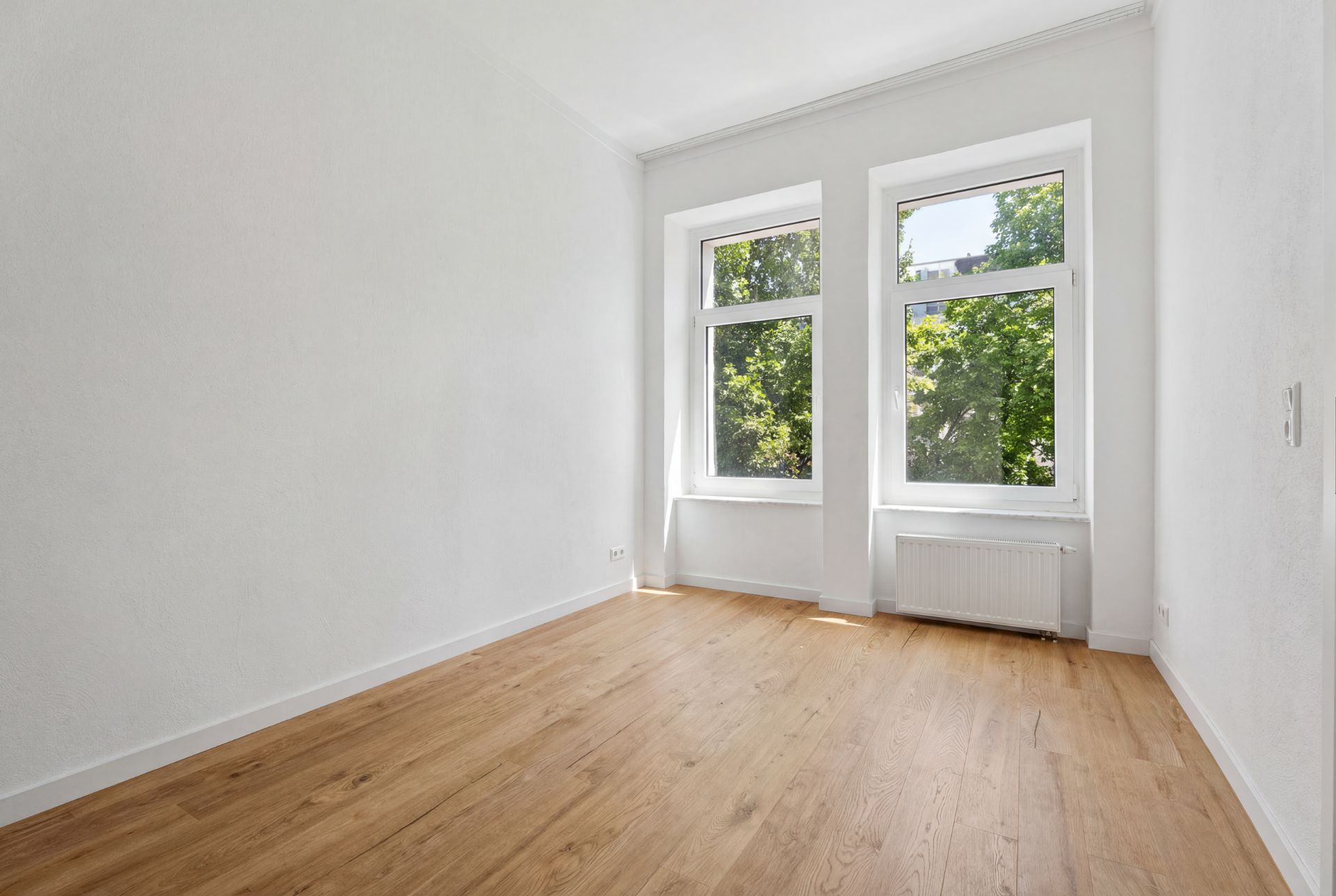 Helles, leeres Zimmer mit zwei großen Fenstern, weißem Heizkörper und Holzfußboden.