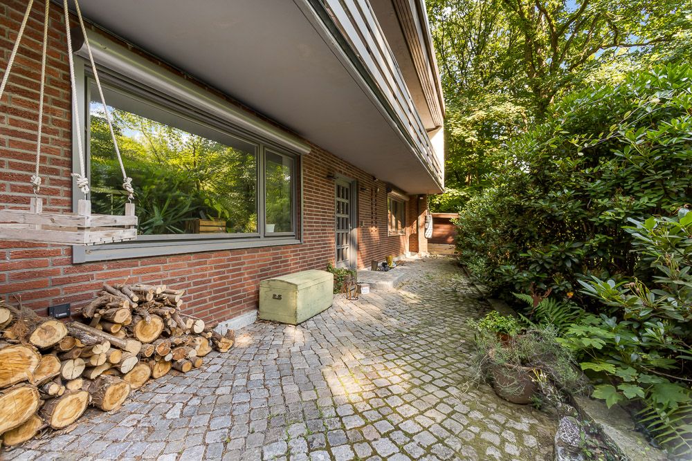 Gepflasterter Weg an einem Backsteinhaus mit Fenster, Holzstapel und viel Grün.