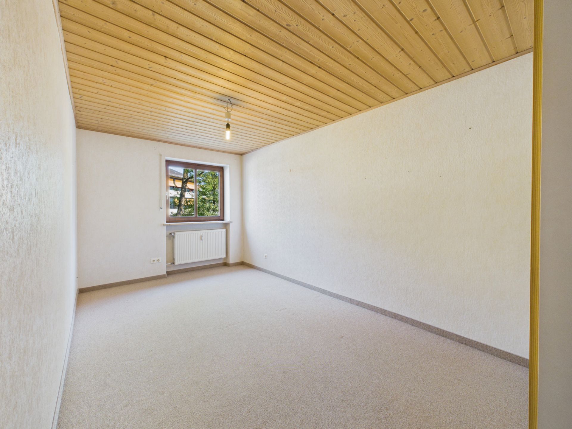 Leerer Raum mit hellem Teppich, Holzdecke und Fenster mit Blick nach draußen.