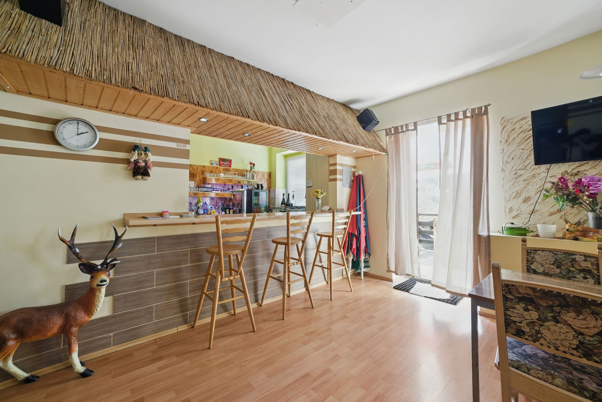 Wohnzimmer mit Bartheke, Holzstühlen, Fernseher, Plastikreh und Uhr an der Wand.