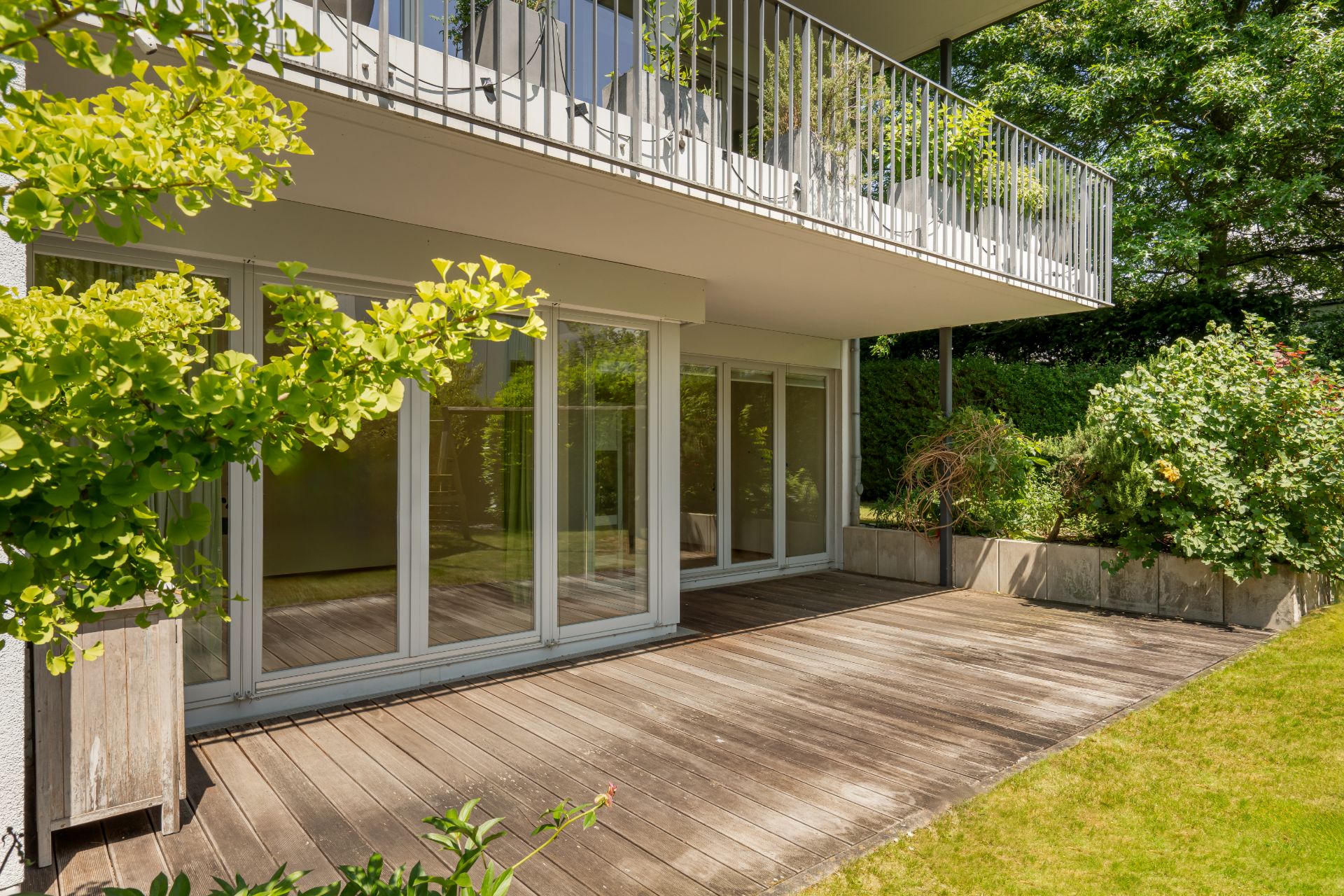 Holzterrasse mit großen Glas-Schiebetüren, begrüntem Garten und Balkon darüber.