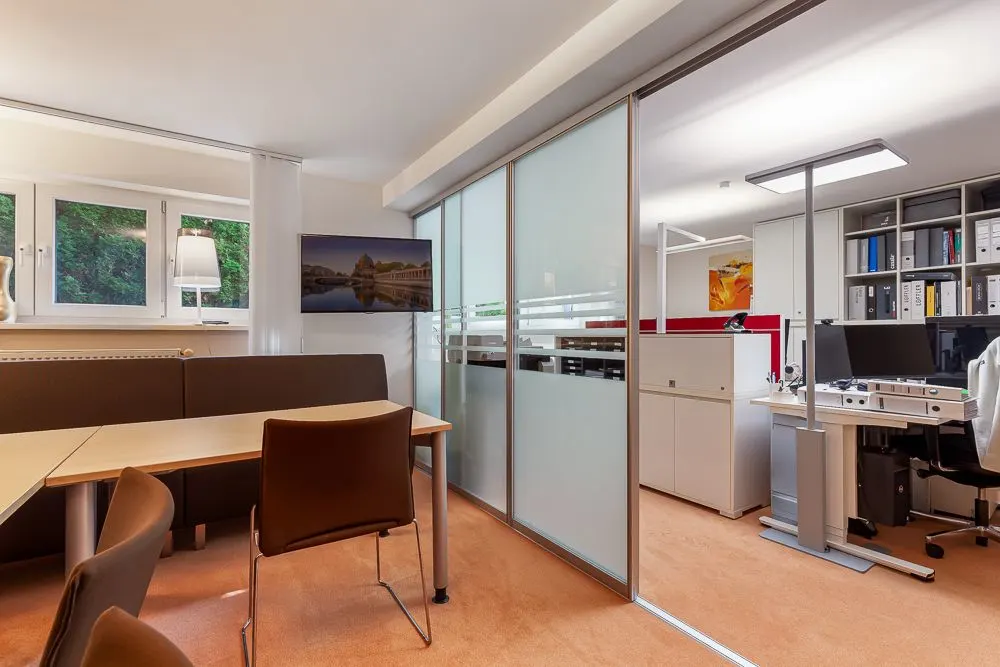 Modernes Arbeitszimmer mit Schreibtischen, Stühlen und Regalen, durch Glaswand getrennt.