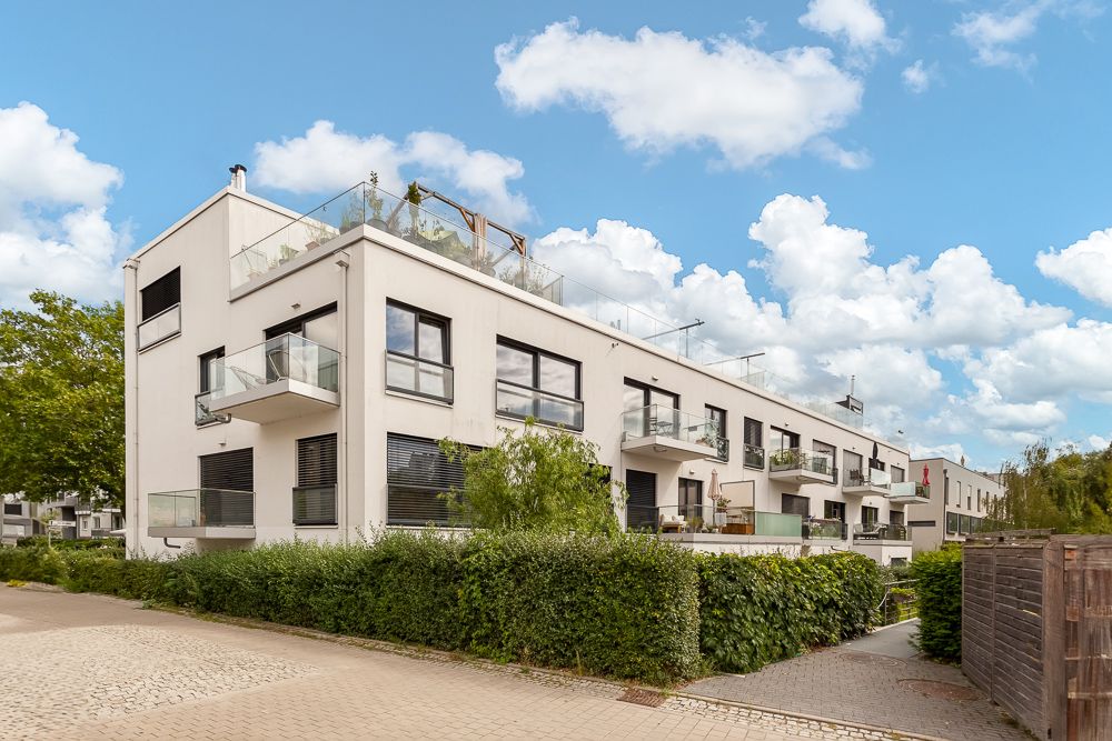Modernes, weißes Wohngebäude mit Balkonen und Dachterrasse, umgeben von Hecken und Gehweg.