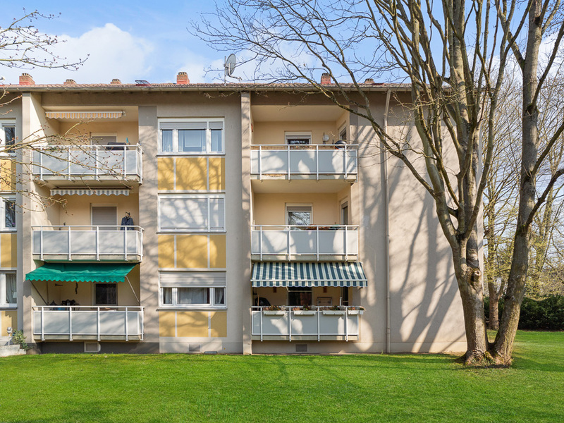 Dreistöckiges Wohnhaus mit Balkonen und Markisen, davor eine Rasenfläche und Bäume.