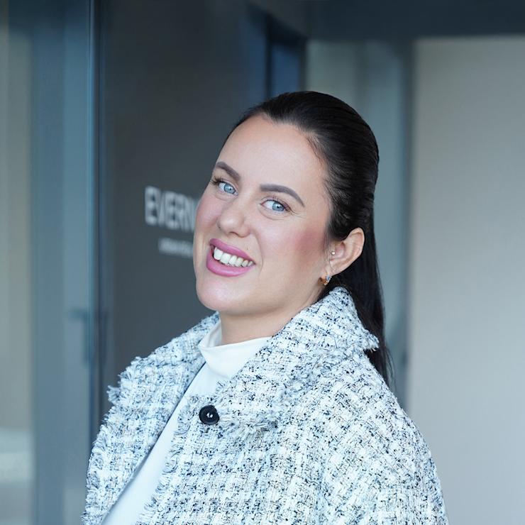 Frau mit hellem Mantel steht lächelnd vor einer Glastür in einem Innenraum.